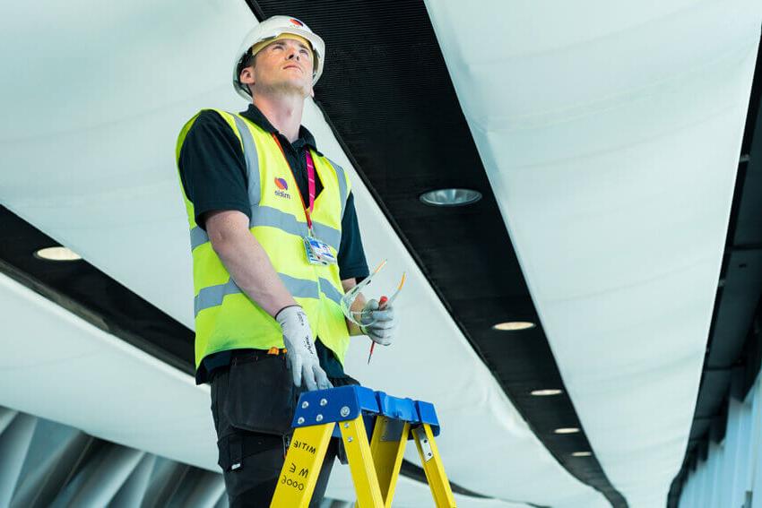 Mitie male engineer fixing a light while standing on a ladder